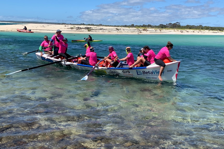 Women wearing pink shirts embrace as they stand next to a surfboat 