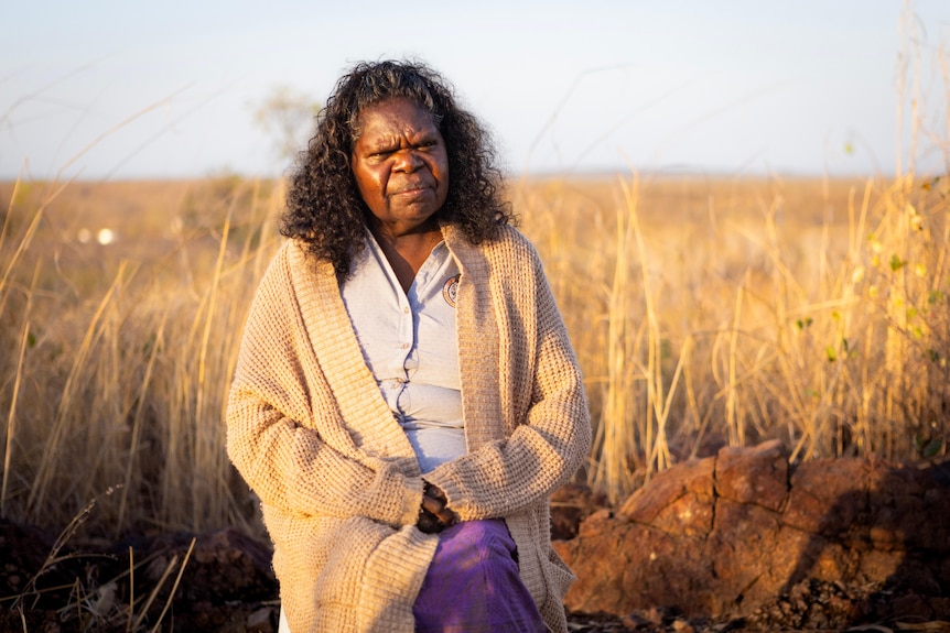 An elderly Indigenous woman standing in an outback savannah, looking behind the camera.