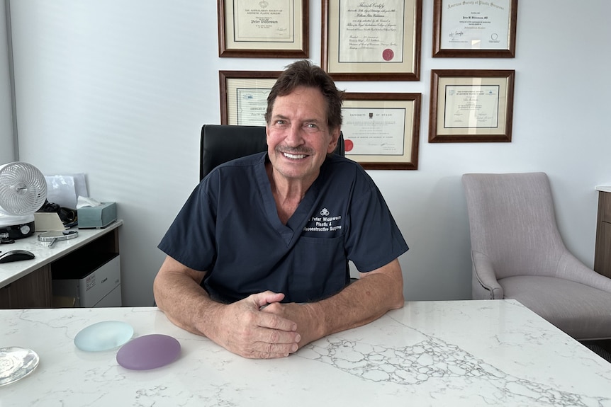 A smiling, dark-haired man with a mustache sits at a desk.  She wears scrubs.