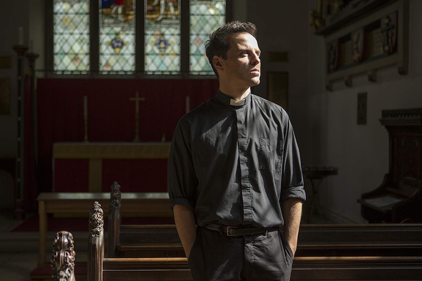 A man in priest blacks with a white colour stares out of frame at light streaming through a church window.