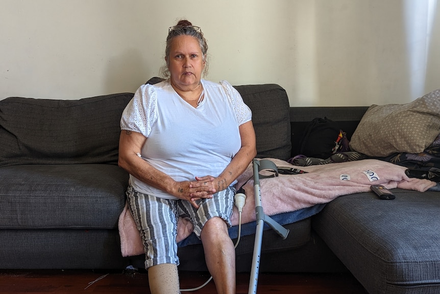Cherie Nannup sits on the couch with a pillow and blanket where she sleeps and her crutch sitting next to her. 