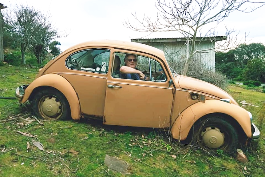 Wide shot from side of old honey brown coloured VW beetle in a paddock, with an older man with sunglasses in front seat.