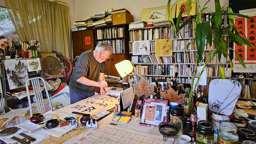 Franz Cheung standing and doing calligraphy in studio filled with books, pens, paintings, paper and ink 