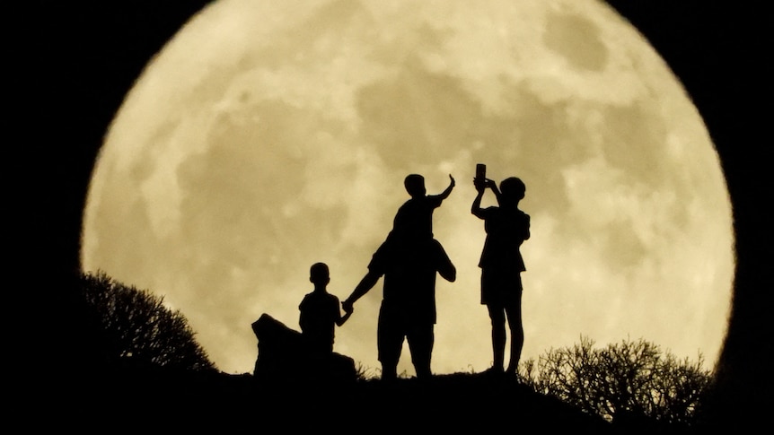 Four people are silhouetted against the backdrop of the moon, which looks massive thanks to a special camera lens