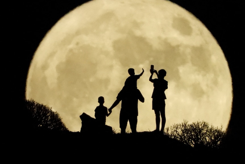 Four people are silhouetted against the backdrop of the moon, which looks massive thanks to a special camera lens