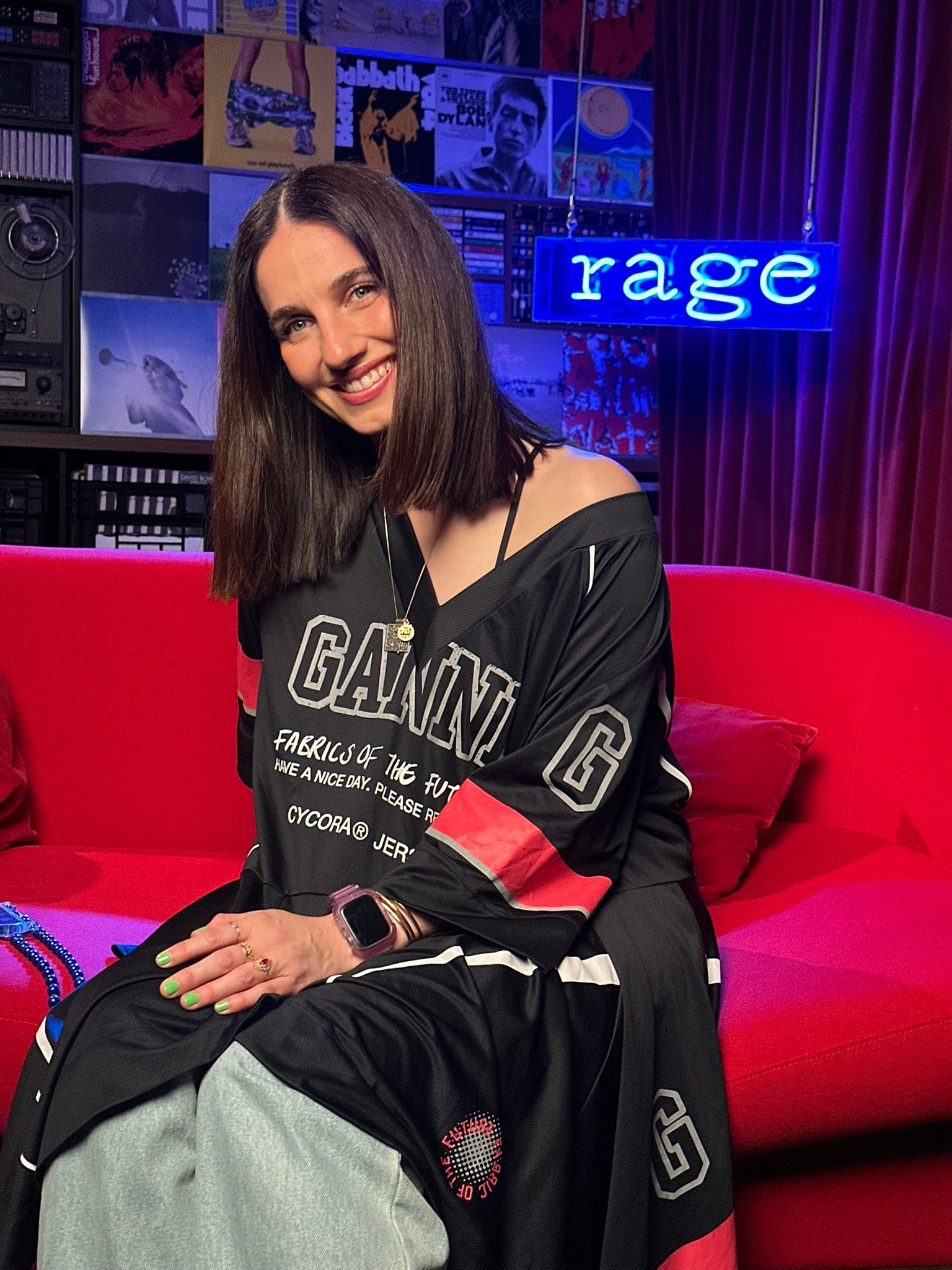 Nina Las Vegas sits smiling on a red couch in front of a blue neon sign that reads rage