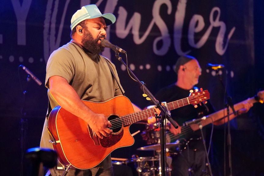 A man wearing a blue cap plays the guitar while singing into a microphone.