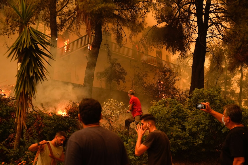 Μια ομάδα ανθρώπων που στέκεται και παρακολουθεί ένα σπίτι που καίγεται από καπνό που πλημμυρίζει τον αέρα γύρω τους.