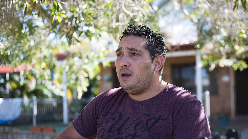 A male singer plays his guitar and is mid song as he stares off to the left of the photo, trees and house in background