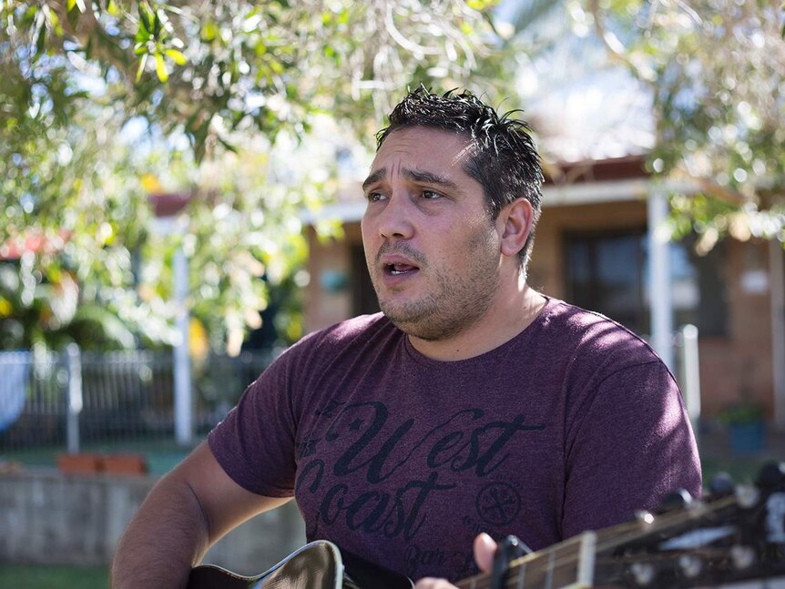 A male singer plays his guitar and is mid song as he stares off to the left of the photo, trees and house in background