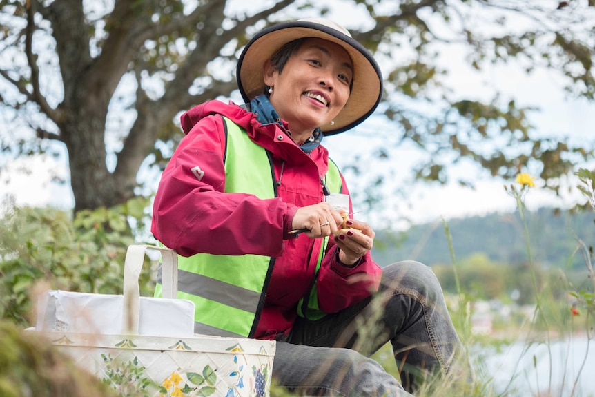 In a leafy green spot outside, a woman wearing wide-brimmed hat and high-vis vest smiles. She cuts something small and yellow.