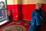 A woman with blonde hair sitting on a red regu with red wall in background.