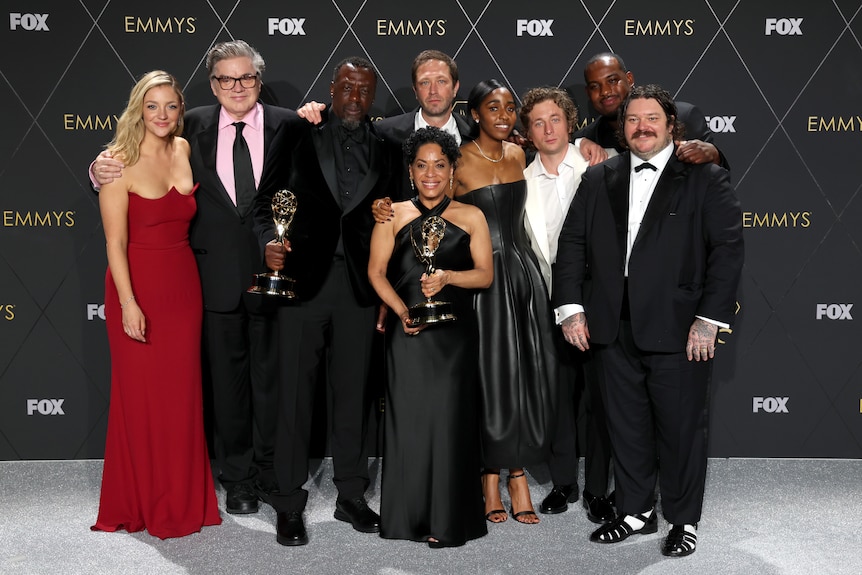 The Bear cast poses together in front of an Emmy Awards poster. Two cast members are holding up their Emmy Awards.