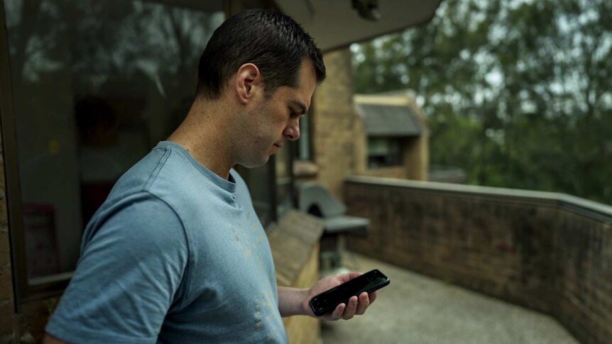 Man with black hair in blue t-shirt stands on balcony looking at phone in his hand with brick building and trees in background.