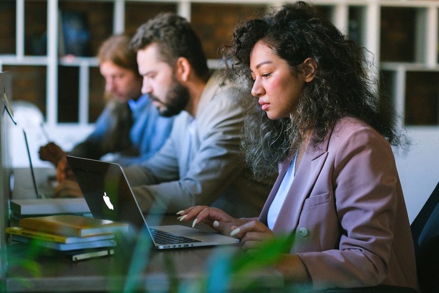 A photo of three people working closely at their computers at a shared desk