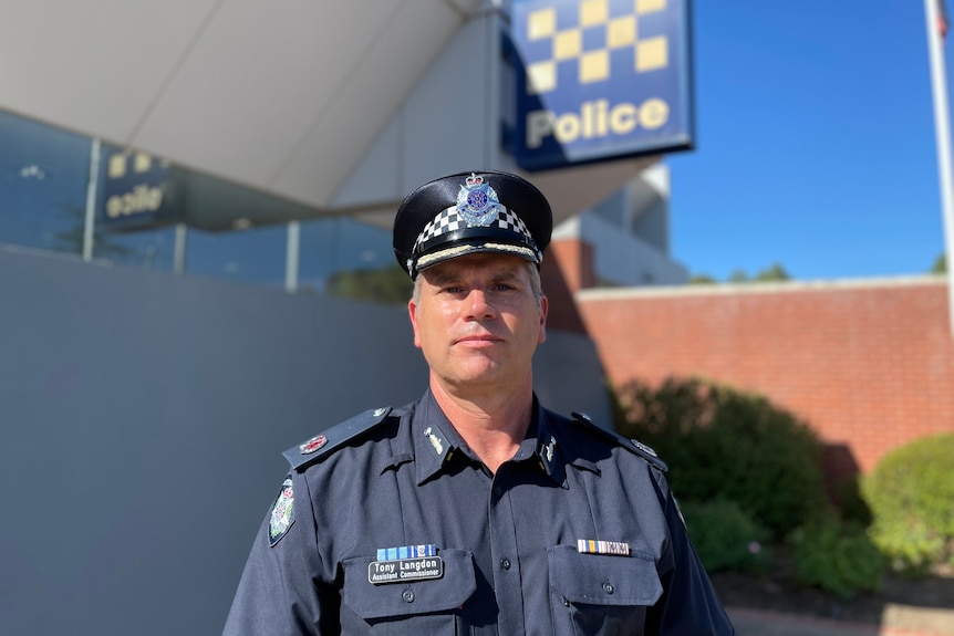 A portrait of a police officer in uniform with a police station and sign behind him,