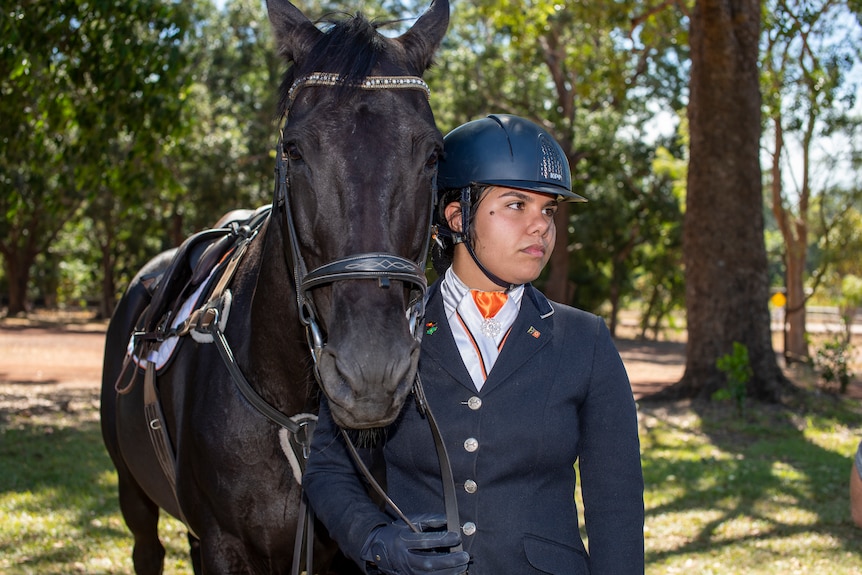 A young Aboriginal woman wears showjumping gear and holds the reins of a black horse. She is looking away from the camera