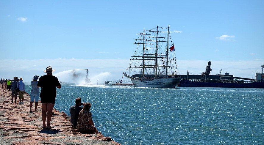 People watch a tall ship from a breakwater looking over a port. 