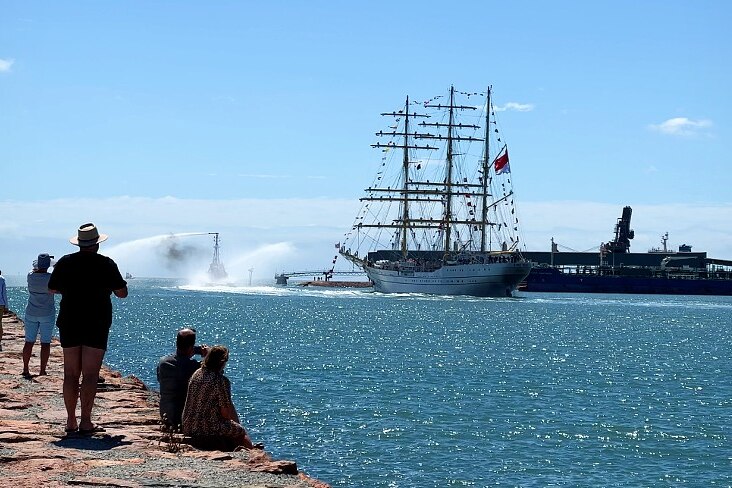 People watch a tall ship from a breakwater looking over a port. 