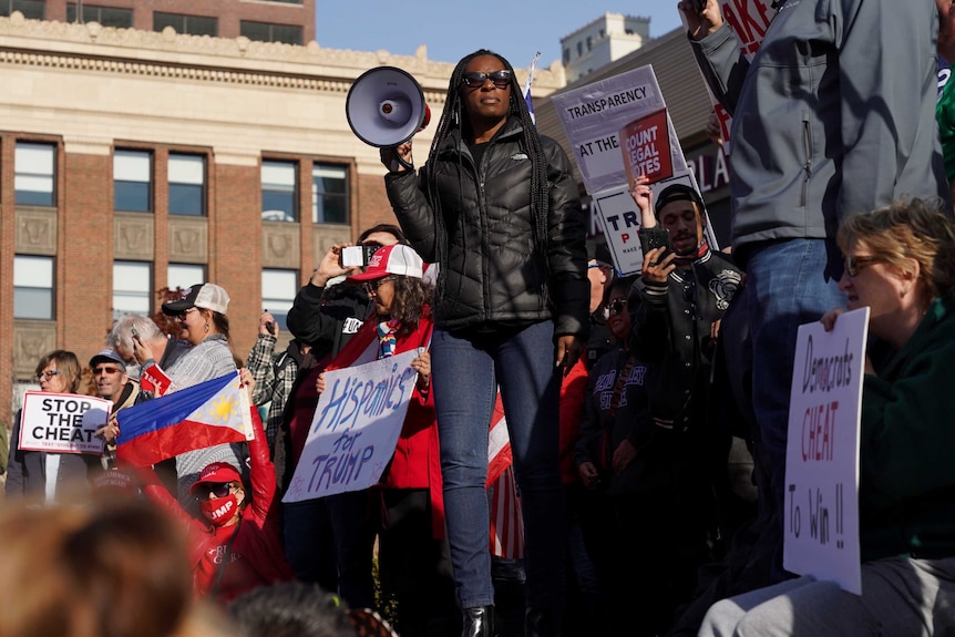 a black woman with a megaphone stands in front of a crowd with pro-Trump placards