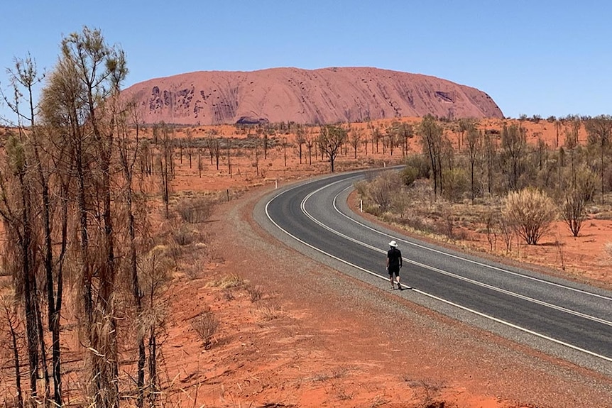 A bitumen road stretches out towards Uluru. A man walks along its edge. 