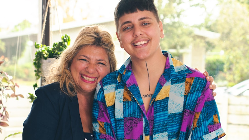 A woman in a dark blazer laughs while hugging a young person in a bright shirt