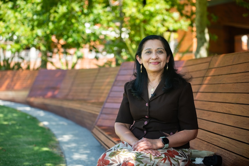 A woman in a brown shirt and flowery skirt sits on a brown bench near some grass and trees.