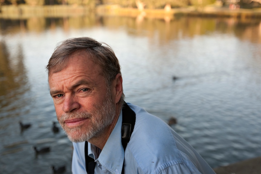 A man with gray hair and a blue shirt sits in front of an outback waterhole looking at the camera.