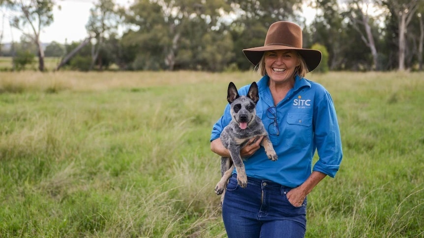 A smiling woman in a blue shirt and brown hat stands in a paddock holding an adorable puppy.