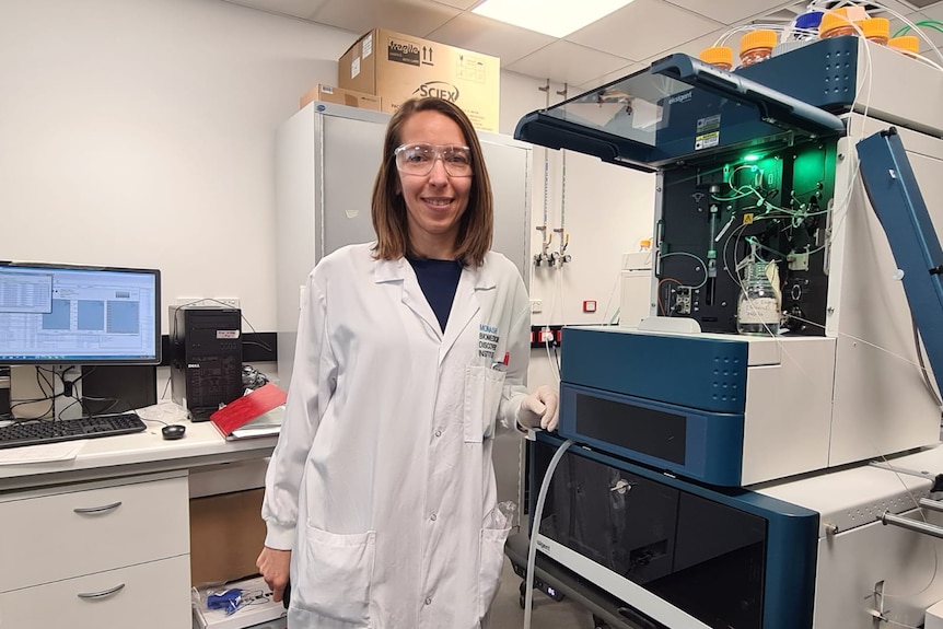 a woman in a white lab coat stands in front of machines.