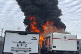 Thick black smoke rises with bright orange flames underneath, coming out of a caravan, the flames touching a caravan beside it.