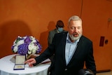 A bearded middle aged white man points at a gold medal in a display case on a table