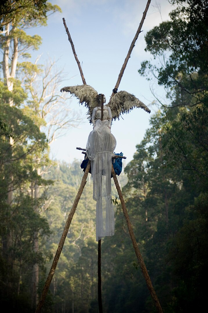 A woman dressed as an angel sits on top of tall wooden poles in a forest