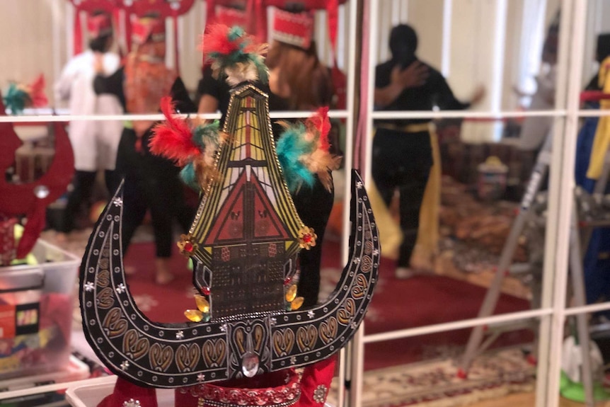An Indonesian headdress sits on a bench in front of a mirror reflecting women dancing in a room.