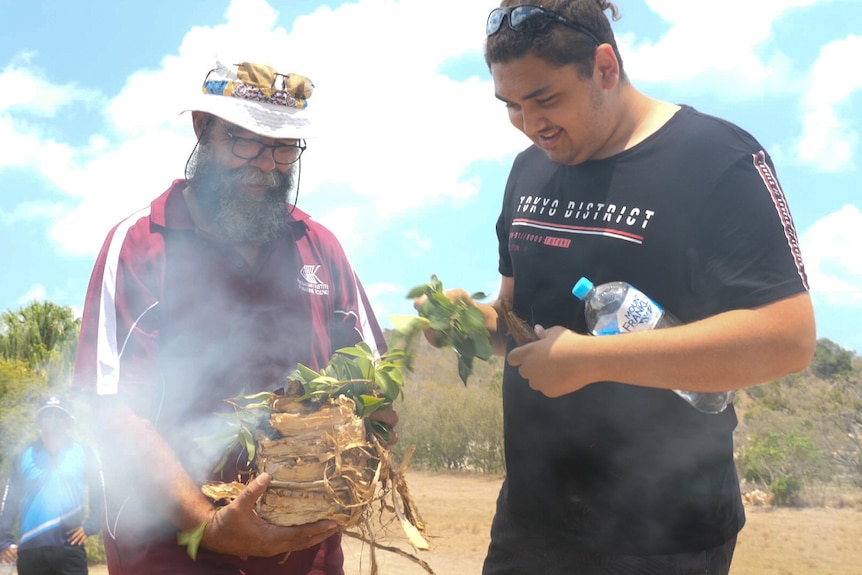 Bob Muir and a young Woppaburra person hold some smoke-filled bark and leaves