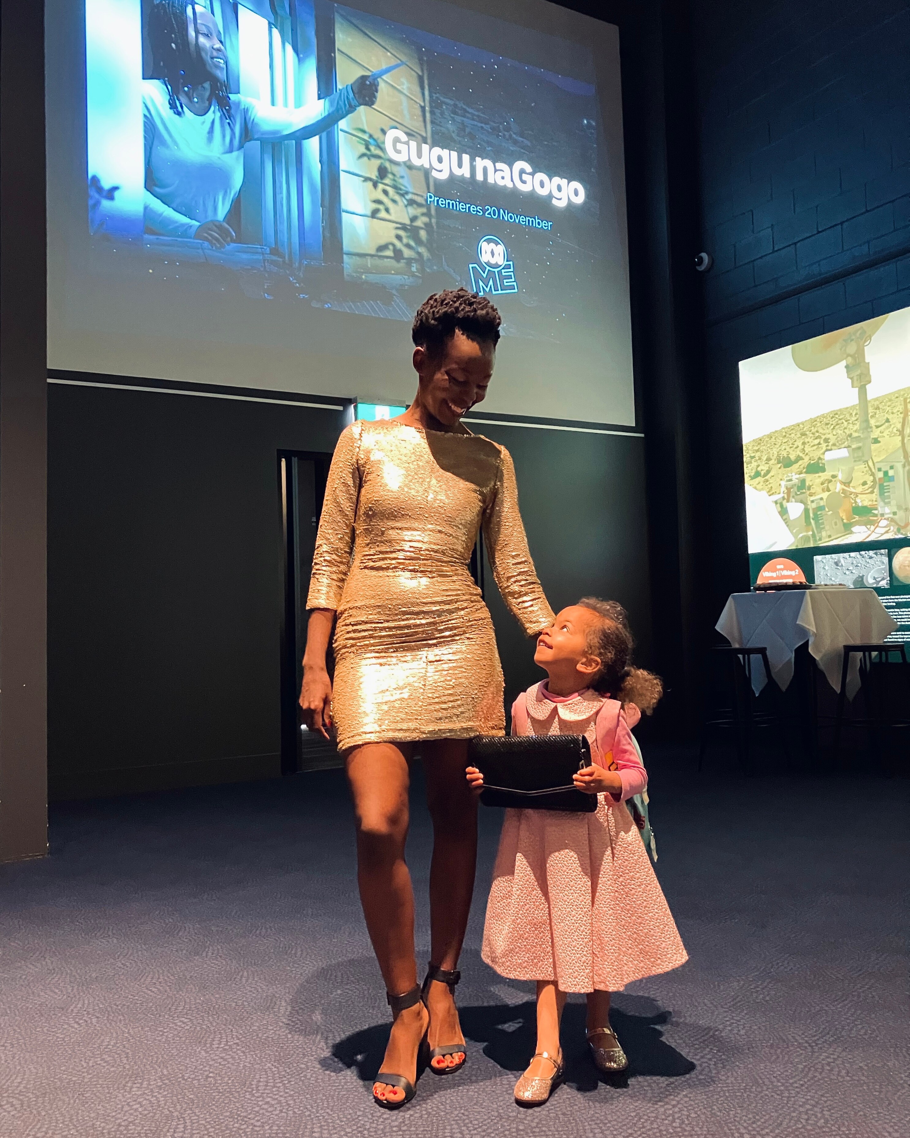 Taku Mbudzi wears a gold dress and stands with her young daughter in a media room at her Gugu naGogo film premiere.