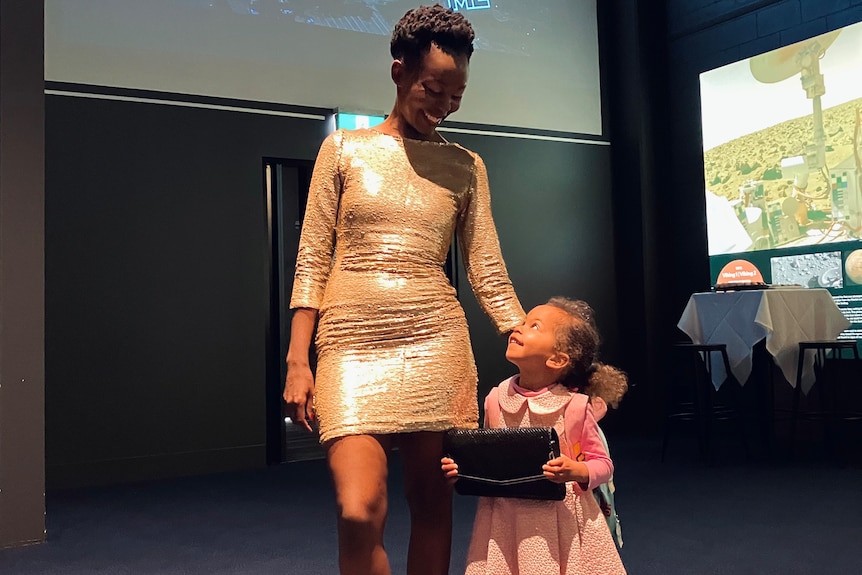 Taku Mbudzi wears a gold dress and stands with her young daughter in a media room at her Gugu naGogo film premiere.