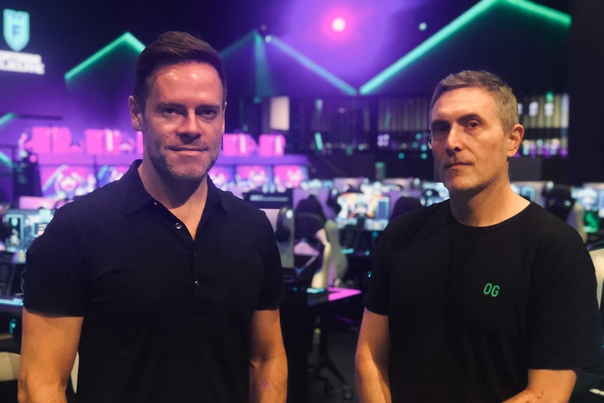 Two men in black t-shirts stand in front of a neon lights.