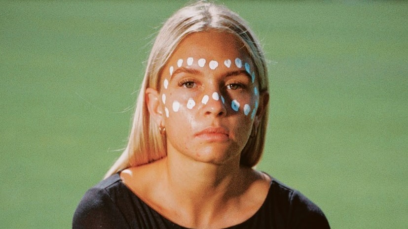 A young Aboriginal woman with traditional facial decorations looks at the camera