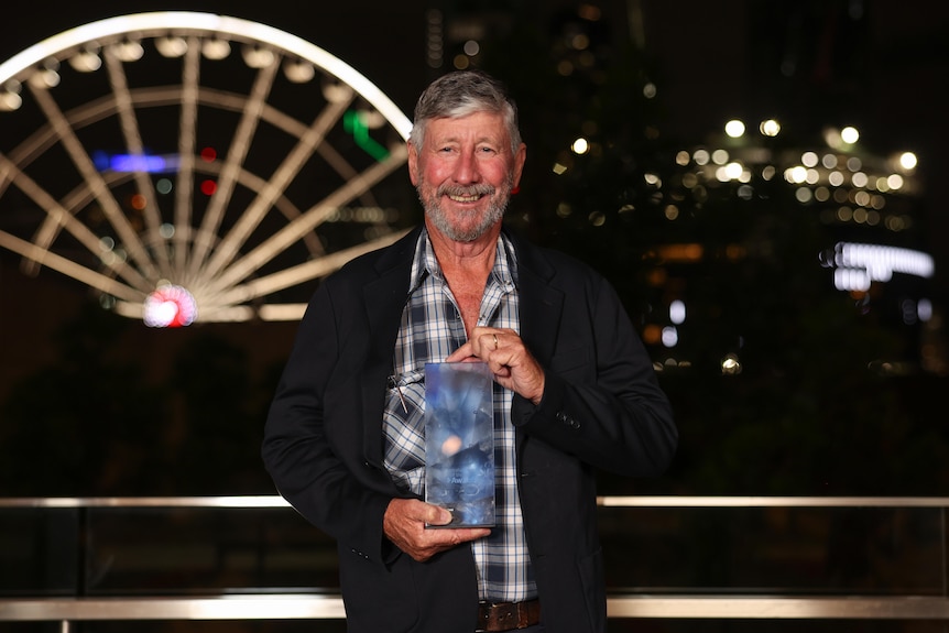 A man in a blue plaid shirt holds a glass award in front of a lit up ferris wheel.