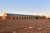 A large stone woolshed with curved roof sits against a bright blue sky at dusk with an old car in the foreground