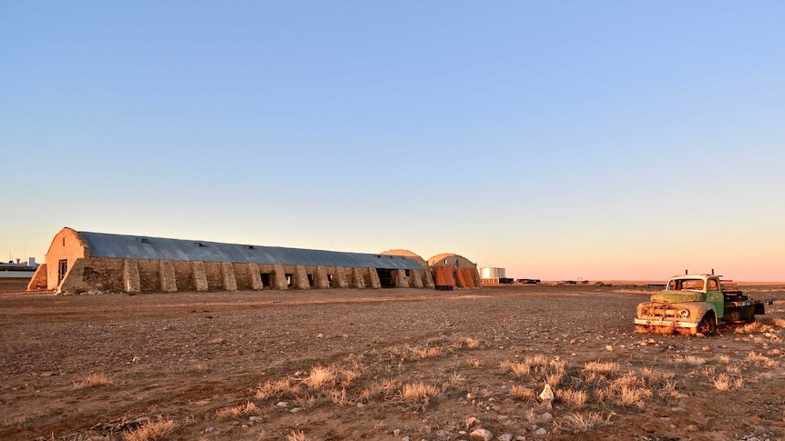 A large stone woolshed with curved roof sits against a bright blue sky at dusk with an old car in the foreground