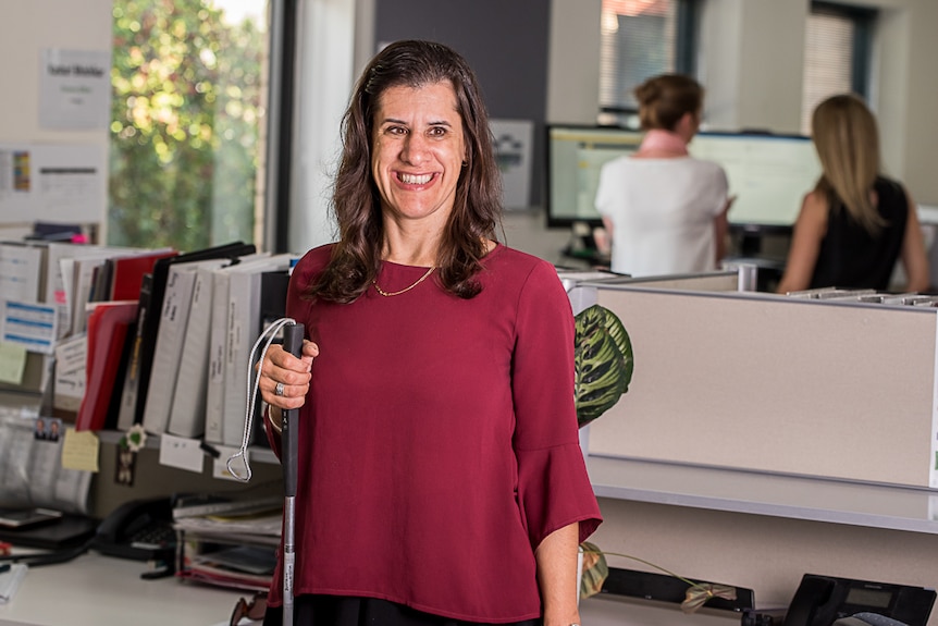 A woman in a corporate office environment, who is blind and holding a cane, smiling and looking past the camera.