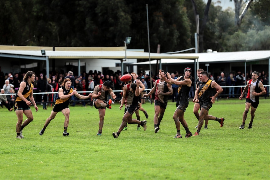 A player kicks a football under pressure as other players and spectators watch on at a country footy ground 