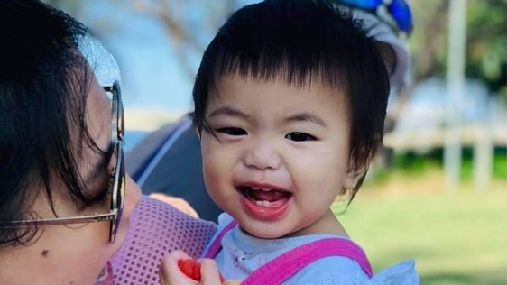 A dark-haired baby girl wearing a blue top and pink rompers over the top being held by her mother who is side on to camera 