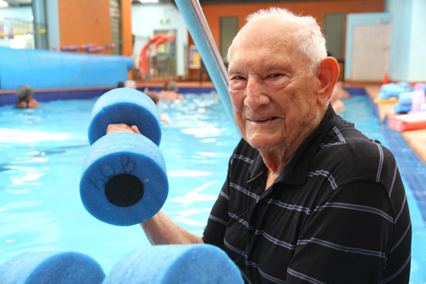 An elderly man grins at the camera as he lifts foam hand weights by the pool side.