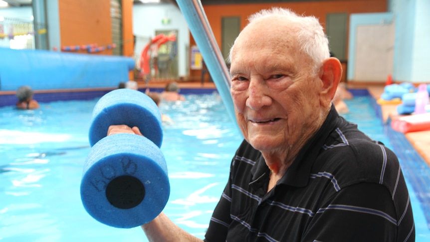 An elderly man grins at the camera as he lifts foam hand weights by the pool side.