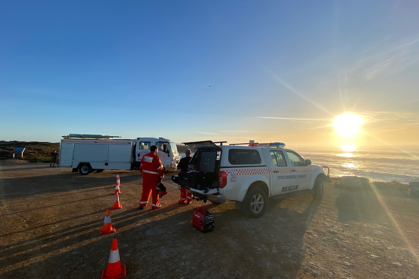 SES workers stand near two SES vehicles parked near the beach as the sun rises over the ocean.