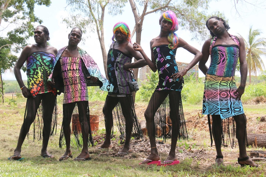 Five trans women in neon printed outfits pose, two in rainbow wigs