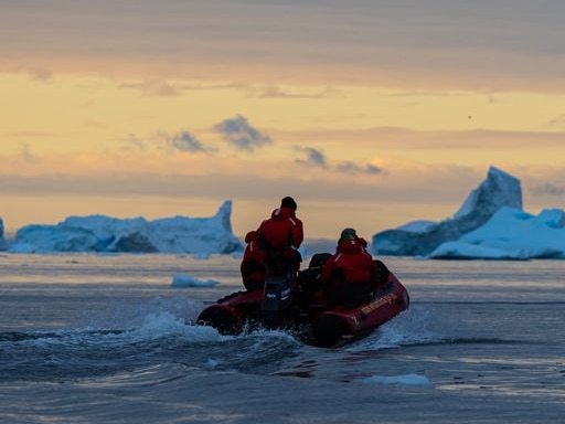Two people in inflatable boat heading through icebergs at sunset